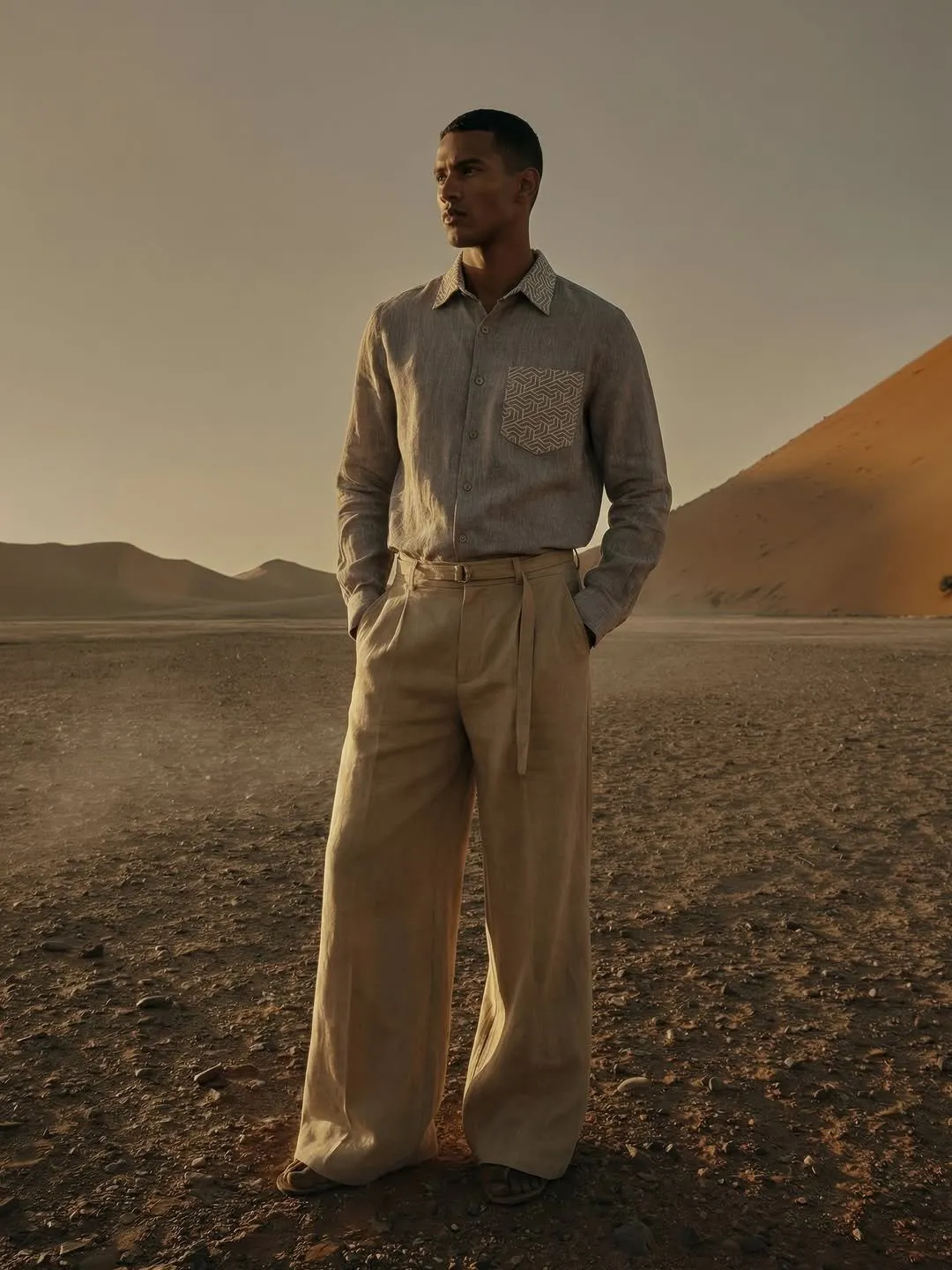 Model in a taupe linen shirt and wide sand linen trousers, standing in the desert at dusk